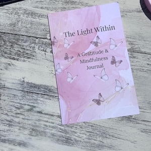 May include: A pink journal titled "The Light Within: A Gratitude & Mindfulness Journal" with butterfly illustrations. The cover has a watercolor effect with gold accents. The journal is on a light-colored wooden surface.