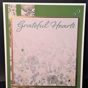 May include: A handmade greeting card with the words "Grateful Hearts" in script. The card features a floral and pumpkin pattern, with green and gold accents, and decorative gems. The card is suitable for fall or Thanksgiving.