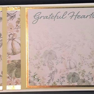 May include: A decorative card with the words "Grateful Hearts" in script. The card features a floral and pumpkin pattern in soft green and pink tones. Gold accents and embellishments add a touch of elegance. The card is framed in a black border.