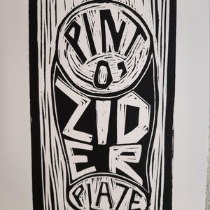 May include: Black and white linocut print featuring the words "PINT OF CIDER" in a stylized design. The artwork has a bold, graphic quality, with a textured, hand-carved appearance. The print is rectangular and set against a white background.