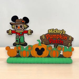 May include: A decorative display featuring a scarecrow with Mickey Mouse ears, a wooden sign reading Mickey's Pumpkin Patch, and several orange pumpkins with green stems and Mickey Mouse ear shapes, all set on a textured green base.