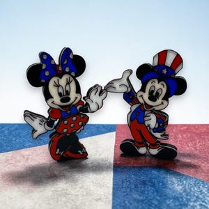 May include: Two cartoon character pins, Minnie Mouse and Mickey Mouse, are depicted in patriotic attire. Minnie Mouse wears a red and white polka dot dress and a blue bow. Mickey Mouse is in a red, white, and blue outfit with a top hat.