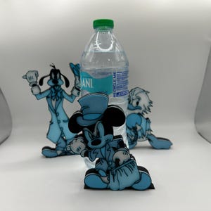 Op de afbeelding: Een doorzichtige waterfles met een groene dop staat achter drie uitgesneden tekenfilmfiguren. Mickey Mouse, Goofy en Donald Duck zijn afgebeeld in lichtblauwe outfits. De figuren staan op een wit oppervlak.