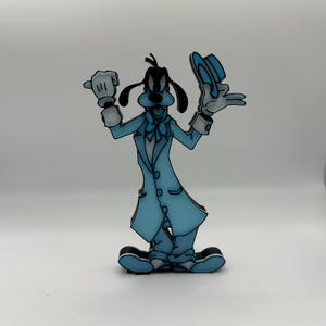 Op de afbeelding: Een lichtblauwe cartoonfiguur van Goofy, een Disney-personage, in een lange jas en hoed. De figuur houdt zijn hoed in &eacute;&eacute;n hand en heeft een handschoen aan de andere hand. De figuur heeft zwarte oren en een zwarte neus.