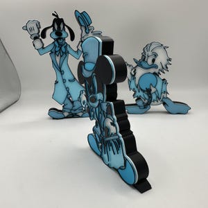 Op de afbeelding: Driedimensionale figuren van Disney-personages Mickey Mouse, Goofy en Donald Duck. De figuren zijn zwart met lichtblauwe accenten. Goofy draagt een blauw pak, Mickey Mouse een blauw outfit en Donald Duck een blauw outfit.
