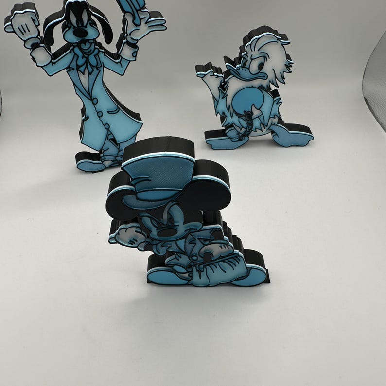 Op de afbeelding: Drie stripfiguurbeeldjes: Goofy, Donald Duck en Mickey Mouse. Elke figuur heeft een lichtblauw en zwart kleurenschema. Goofy staat met opgeheven armen. Donald Duck staat in een vergelijkbare pose. Mickey Mouse knielt.