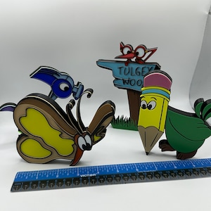 May include: A set of four colorful, hand-painted wooden figurines. The figurines include a butterfly, a pencil, a bird, and a sign that reads "Tulgey Wood".