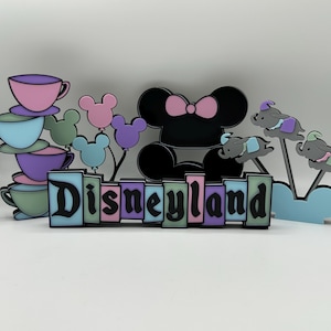 May include: A pastel-colored Disneyland sign with Mickey Mouse ears, balloons, a teacup stack, and Dumbo the Flying Elephant. The sign is made of wood and has a black outline.