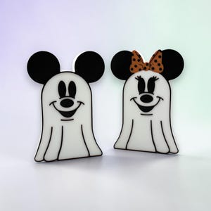 May include: Two flat, white ghost-shaped figures with black mouse ears. The left figure has a simple smiling face. The right figure has a smiling face with eyelashes and an orange bow with black polka dots on top of its head. Both are white.
