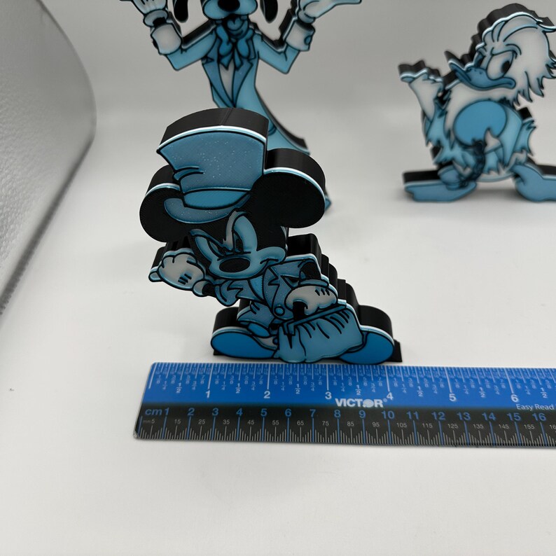 Op de afbeelding: Blauwe en zwarte Mickey Mouse, Goofy en Donald Duck beeldjes. Een blauwe liniaal met centimeter maten staat op de voorgrond. Het Mickey Mouse beeldje is ongeveer 7 cm hoog.