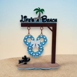 May include: A decorative sign with the text "Life's a Beach" features a Mickey Mouse-shaped design with blue wreaths and a white bow. A palm tree and a silhouette of Mickey Mouse are also included in the design. The sign is brown and stands on a sandy base.