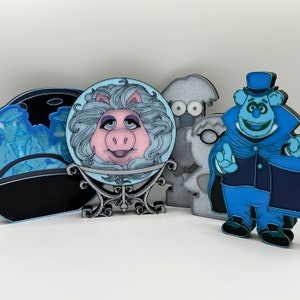 May include: Five enamel pins featuring characters from the Muppets. The pins are blue, white, and black. The pins are of Miss Piggy, a ghost, a monster, and two other Muppets.