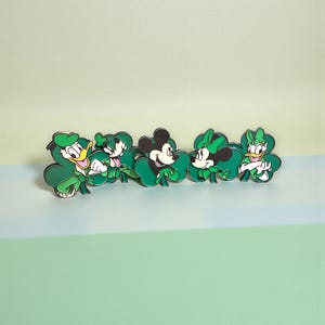 May include: A row of five green character pins, each set on a shamrock. The characters include Donald Duck, Goofy, Mickey Mouse, Minnie Mouse, and Daisy Duck. The pins are set against a light green and blue background.