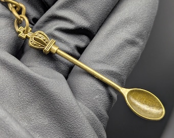 May include: A small, antique-style spoon pendant with a crown design. The spoon is made of a gold-colored metal and has an oval bowl. The crown is detailed with small beads and a cross on top. The spoon is attached to a chain.