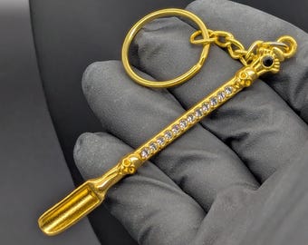 May include: A gold-colored keychain featuring a decorative wand-shaped charm. The charm is adorned with clear crystals and a black gem at the top. The wand has a scoop at the end and is attached to a gold ring and chain.