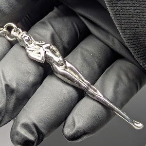 May include: A silver-colored pendant in the shape of a slender female figure, with a chain loop at the top. The figure is detailed with smooth curves and a polished finish. The pendant is held against a dark background.