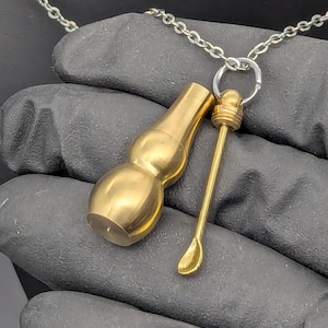 May include: A gold-colored pendant necklace featuring a small, gourd-shaped container and a slender spoon-like tool. The pendant hangs from a silver chain. The items are made of a shiny, metallic material.