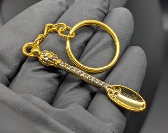 May include: A gold-colored spoon-shaped keychain with a ring and chain. The spoon is adorned with small, clear crystals along the handle and on the decorative top. The keychain is held against a dark background.