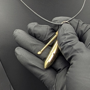 May include: A gold-colored necklace with a pendant shaped like a small, thin, pointed object. The pendant has a small, round hole at the top for the necklace to pass through. The pendant is being held by a gloved hand.