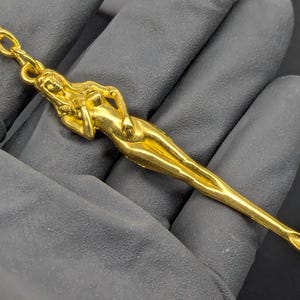 May include: A gold-colored keychain featuring a stylized reclining female figure. The figure is elongated and detailed, with a chain and clasp at the top. The keychain is made of a shiny, metallic material.