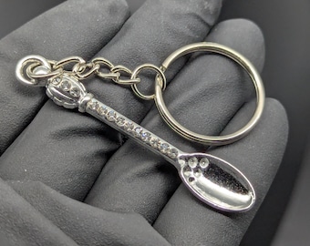 May include: Silver spoon keychain with clear crystal accents. The spoon features a decorative handle and a chain that connects to a large silver ring. The spoon's bowl has several small holes.