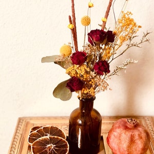 Pode incluir: Arranjo de flores secas em uma garrafa de vidro marrom com uma romã vermelha e fatias de laranja secas em um livro com a palavra "ART" e o texto "THE DEFINITIVE VISUAL GUIDE" na capa.