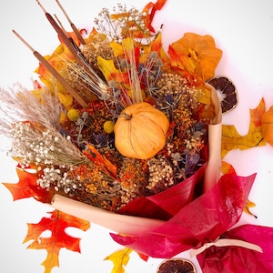 May include: A fall-themed bouquet with dried flowers, leaves, and a small orange pumpkin. The bouquet is wrapped in brown paper and tied with a red ribbon. The bouquet is surrounded by dried orange slices and leaves.