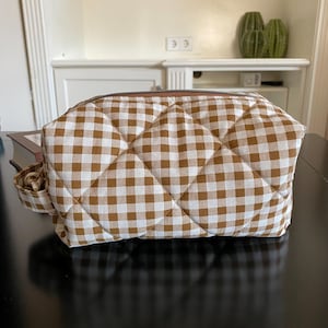 Può includere: Un beauty case trapuntato con un motivo a quadri marrone e bianco. La borsa ha una forma rettangolare con chiusura a zip e un piccolo manico laterale. La borsa è appoggiata su una superficie scura.