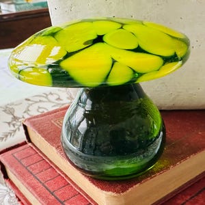 May include: A decorative glass mushroom with a green stem and a yellow and green cap. The mushroom is sitting on top of two antique books with red covers. The mushroom is a unique decorative item.