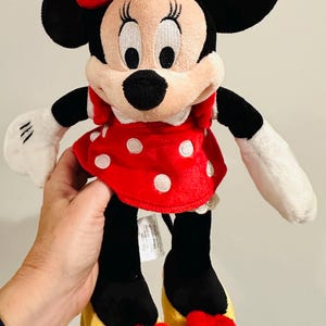 Puede incluir: Mu&ntilde;eca de peluche de Minnie Mouse. Tiene un vestido rojo con lunares blancos, un lazo a juego y zapatos amarillos con lazos rojos. La mu&ntilde;eca tiene orejas negras, nariz negra y guantes blancos. Un personaje cl&aacute;sico de Disney.