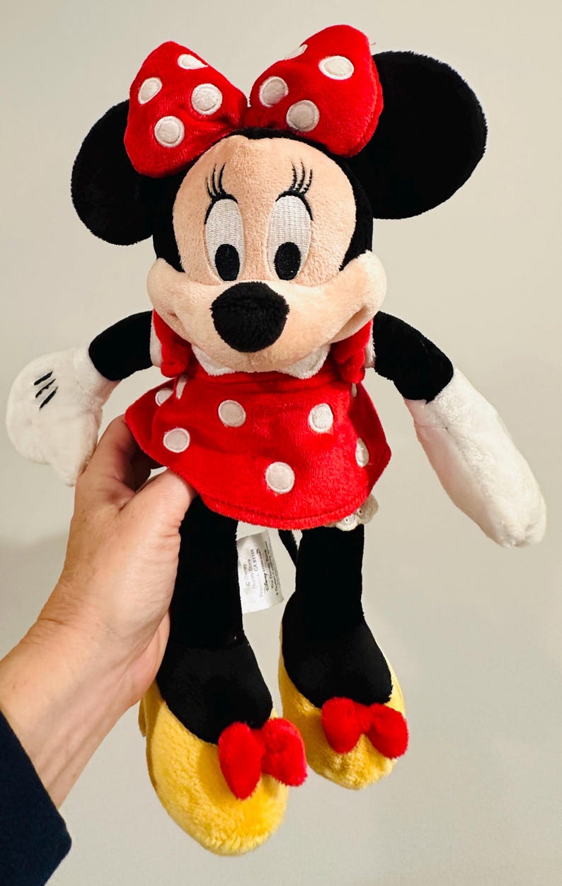 Puede incluir: Mu&ntilde;eca de peluche de Minnie Mouse. Tiene un vestido rojo con lunares blancos, un lazo a juego y zapatos amarillos con lazos rojos. La mu&ntilde;eca tiene orejas negras, nariz negra y guantes blancos. Un personaje cl&aacute;sico de Disney.