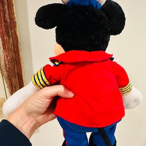 May include: A plush Mickey Mouse doll wearing a red jacket with yellow and blue accents, blue pants, and a white sailor hat. The doll has black ears, a black tail, and white gloves. The back of the doll is visible.