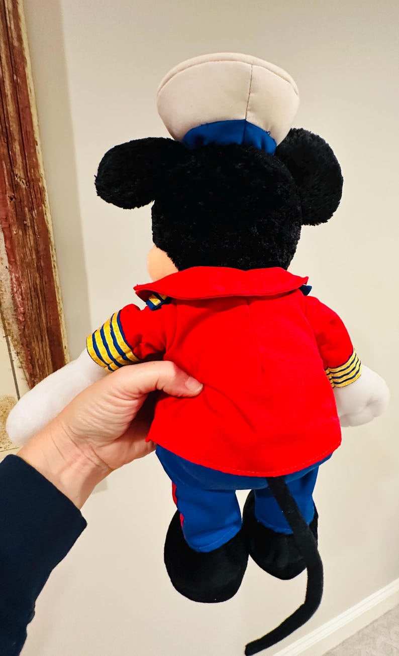 May include: A plush Mickey Mouse doll wearing a red jacket with yellow and blue accents, blue pants, and a white sailor hat. The doll has black ears, a black tail, and white gloves. The back of the doll is visible.