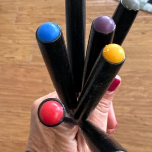 May include: A collection of black, cylindrical objects with colorful, rounded tops. The tops are in various colors, including red, blue, green, yellow, purple, and gray. The objects are held in a hand, with a wooden surface in the background.