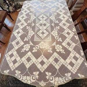 May include: A rectangular cream-colored lace tablecloth featuring an intricate openwork pattern. The design includes a central geometric motif surrounded by floral arrangements and a repeating diamond grid border that extends to the edges.