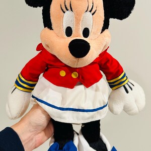 May include: A plush Minnie Mouse doll wearing a red jacket with gold buttons, a white skirt with a blue trim, and white shoes with blue bows. The doll has black ears and a red bow on her head.