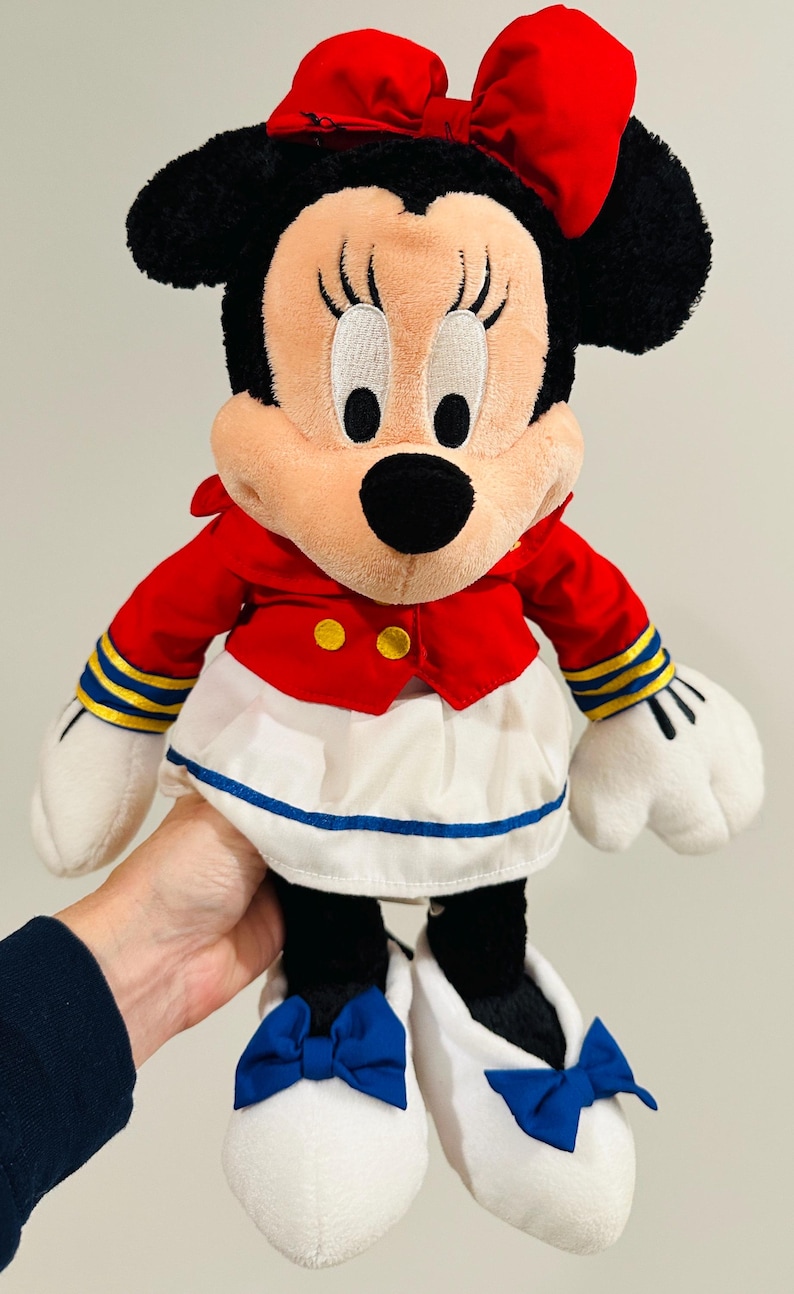May include: A plush Minnie Mouse doll wearing a red jacket with gold buttons, a white skirt with a blue trim, and white shoes with blue bows. The doll has black ears and a red bow on her head.