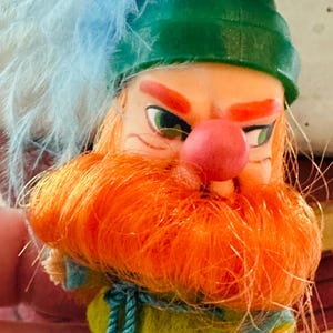 May include: A vintage figurine of a leprechaun with an orange beard and eyebrows, a red nose, and green eyes. He wears a green hat with blue feathers, a yellow jacket, and blue pants. The figurine is held by a hand.