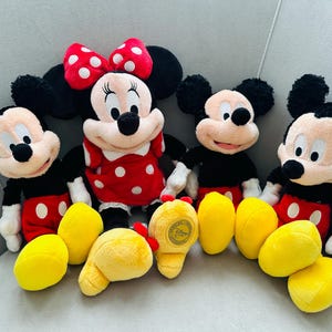 Puede incluir: Cuatro peluches de personajes de Disney: Mickey Mouse y Minnie Mouse. Mickey lleva pantalones cortos rojos, camisa negra y guantes blancos. Minnie lleva un vestido rojo con lunares blancos y un lazo rojo. Pies de peluche amarillos al frente.