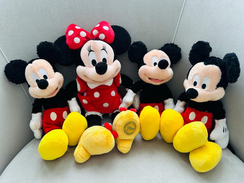 Puede incluir: Cuatro peluches de personajes de Disney: Mickey Mouse y Minnie Mouse. Mickey lleva pantalones cortos rojos, camisa negra y guantes blancos. Minnie lleva un vestido rojo con lunares blancos y un lazo rojo. Pies de peluche amarillos al frente.