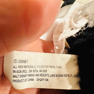 May include: A close-up of a Disney product tag. The tag is white with black text, listing materials, location, and the Disney logo. The tag is held by a hand, with a portion of a black and white plush toy visible.