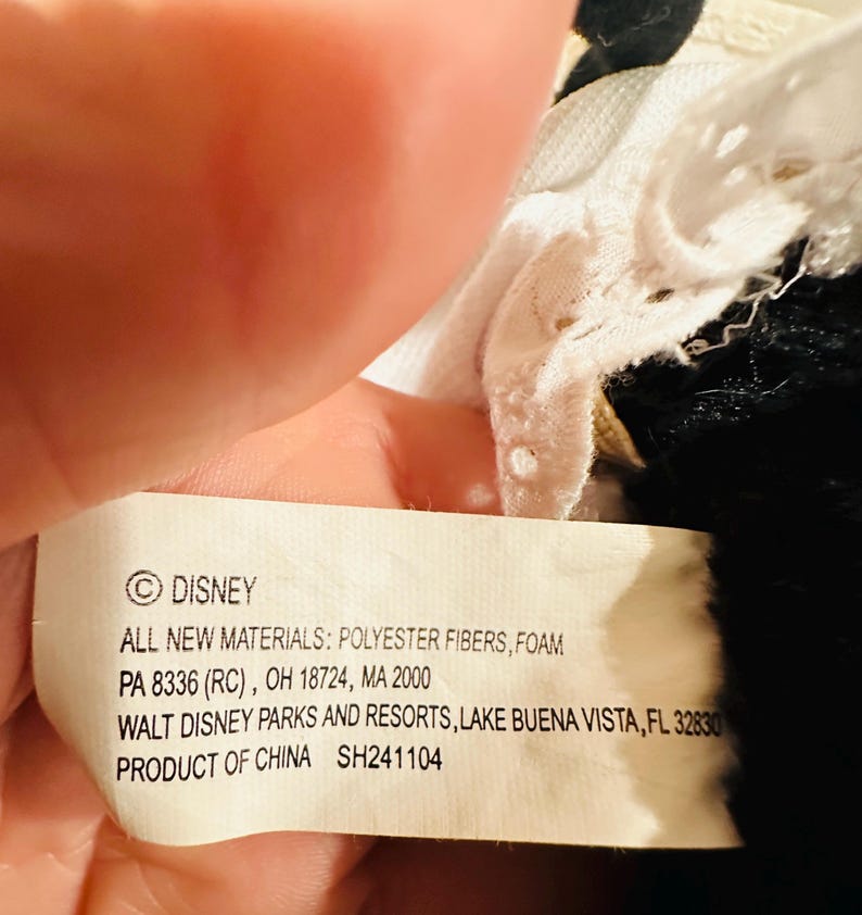 May include: A close-up of a Disney product tag. The tag is white with black text, listing materials, location, and the Disney logo. The tag is held by a hand, with a portion of a black and white plush toy visible.