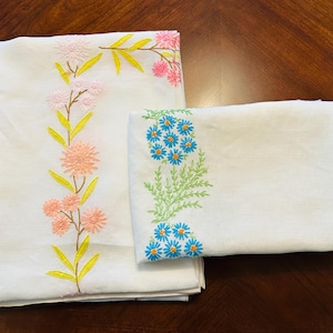 May include: White embroidered bedding set featuring floral designs. The larger piece has a vertical arrangement of pink and peach flowers with yellow leaves. The smaller piece has blue flowers with green leaves. The set is made of a soft, white fabric.