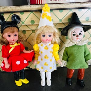May include: Three vintage dolls: a Minnie Mouse doll in a red dress with white polka dots, a doll in a white dress with yellow floral accents and a pointed hat, and a Wizard of Oz doll in a green top and black hat.
