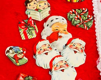 May include: A collection of vintage Christmas-themed paper cutouts on a red background. The cutouts include Santa Claus faces, children singing carols, wrapped gifts, candy canes, bells, and candle decorations. The overall theme is festive and nostalgic.
