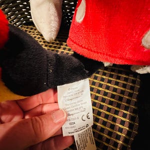 Puede incluir: Primer plano de un peluche de Mickey Mouse. El juguete tiene un sombrero rojo con lunares blancos, pelaje negro y un guante blanco. Una etiqueta con el logotipo y la direcci&oacute;n de Disney Store es visible.