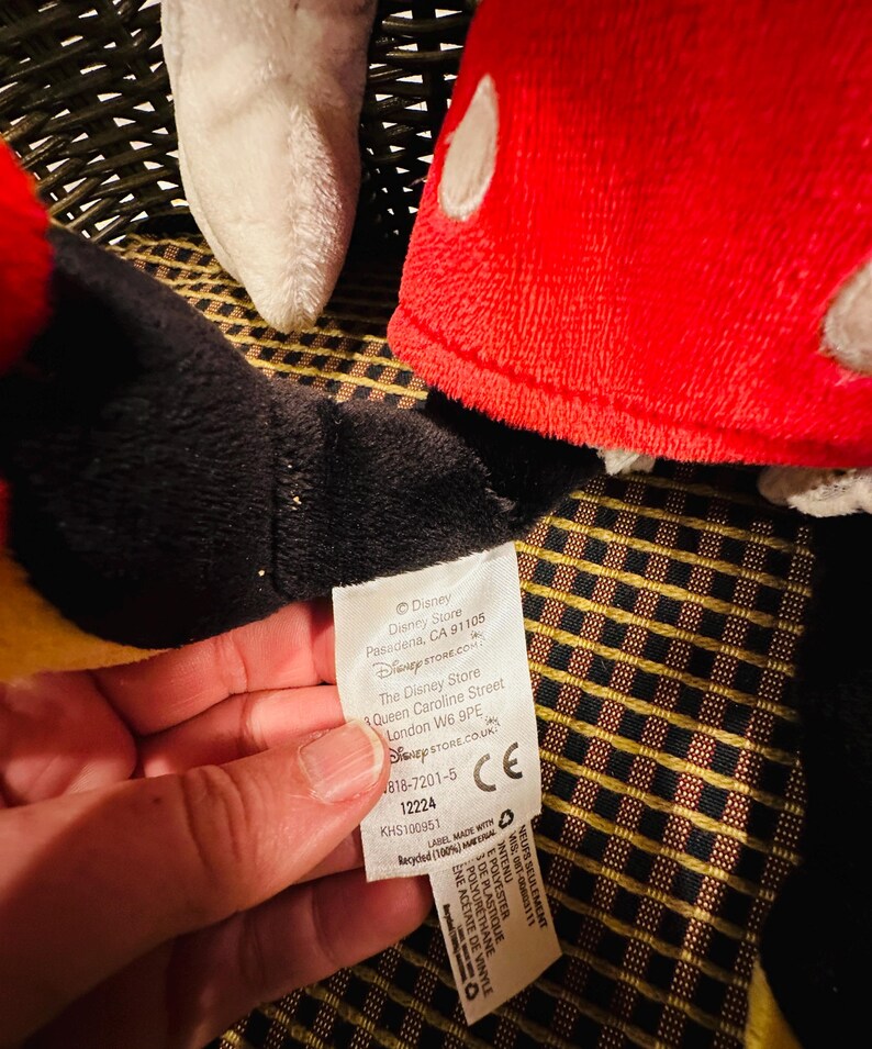 Puede incluir: Primer plano de un peluche de Mickey Mouse. El juguete tiene un sombrero rojo con lunares blancos, pelaje negro y un guante blanco. Una etiqueta con el logotipo y la direcci&oacute;n de Disney Store es visible.
