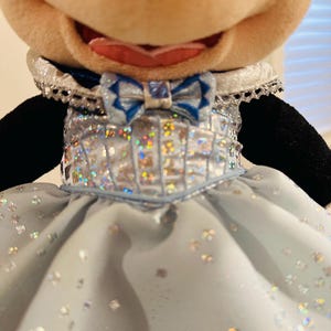 K&ouml;nnte beinhalten: Pl&uuml;schtier einer Figur mit einem gro&szlig;en, l&auml;chelnden Gesicht. Die Figur tr&auml;gt ein hellblaues Kleid mit silbernen Akzenten und einer passenden Fliege. Das Kleid hat ein funkelndes, schillerndes Aussehen, und der Kragen ist mit silberner Spitze verziert.