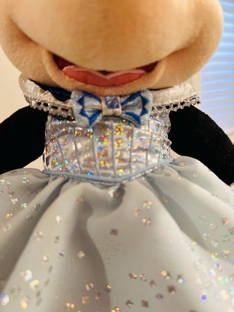 K&ouml;nnte beinhalten: Pl&uuml;schtier einer Figur mit einem gro&szlig;en, l&auml;chelnden Gesicht. Die Figur tr&auml;gt ein hellblaues Kleid mit silbernen Akzenten und einer passenden Fliege. Das Kleid hat ein funkelndes, schillerndes Aussehen, und der Kragen ist mit silberner Spitze verziert.