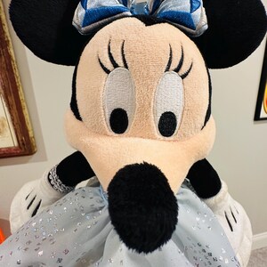 K&ouml;nnte beinhalten: Ein Pl&uuml;sch Minnie Mouse Puppe mit schwarzen Ohren, einem hellrosa Gesicht und einer schwarzen Nase. Sie tr&auml;gt ein hellblaues Kleid mit silbernen Glitzer und einer blau-silbernen Schleife. Ihre wei&szlig;en Handschuhe und schwarzen Arme sind sichtbar.